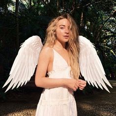 a woman with long blonde hair and white angel wings on her shoulders, standing in front of trees