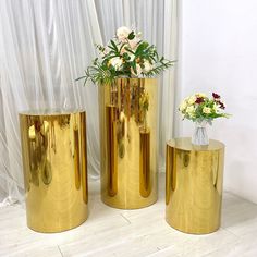 three gold vases with flowers in them sitting on a white floor next to a curtain