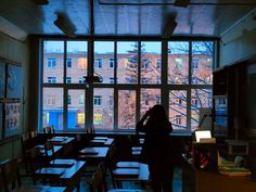 a person standing in front of a large window next to desks and bookshelves