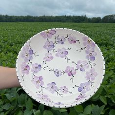 a hand holding a plate with flowers painted on it in the middle of a field