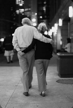 an older man and woman walking down the street at night with their backs to each other