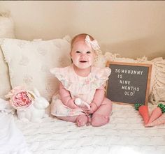 a baby sitting on a bed next to stuffed animals and a sign that says she is nine months old