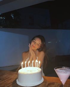 a woman sitting in front of a white cake with lit candles on it next to a drink