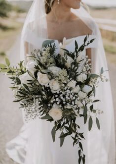 a woman in a wedding dress holding a bridal bouquet with white flowers and greenery