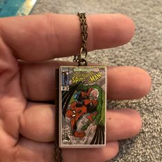 a hand holding a small square pendant with an image of spider - man on it