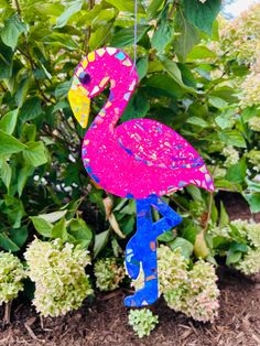 a pink flamingo bird hanging from a tree in front of some green bushes and flowers