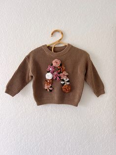 a brown sweater with flowers on it hangs from a hook against a white wall,