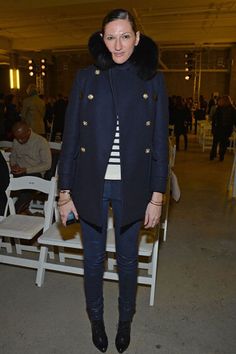 a woman standing in front of white chairs wearing a coat and black pants with fur collar