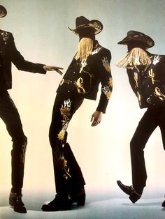 three men dressed in black and gold with long blonde hair, one wearing a cowboy hat