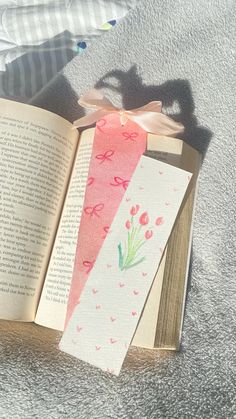 an open book sitting on top of a bed next to a pink flower and ribbon