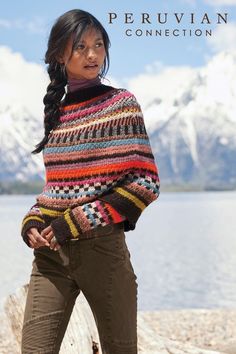 a woman wearing a multicolored sweater standing by the water with mountains in the background