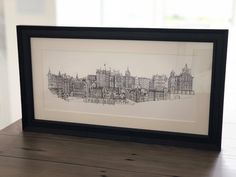 a black and white drawing of a cityscape in a frame on a table