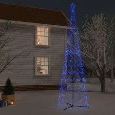 a blue christmas tree in front of a white house with snow on the ground at night