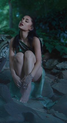 a woman sitting on top of a rock next to a forest filled with green leaves