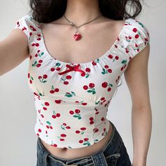 A thin, light-skinned woman's torso. She is wearing a white blouse with small red cherries printed on it. At the top of the blouse is a small red bow. She wears a red, heart-shaped lock necklace and dark jeans. The ends of her dark brown, wavy hair are visible. Short Paragraph, Fresh Cherry, Girl Jacket, Bodycon Dresses Casual, White Short Dress, American Girls, Workwear Fashion, Cherry Print, Crop Top Blouse