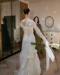 a woman in a white wedding dress is holding flowers and looking at another woman in a green dress