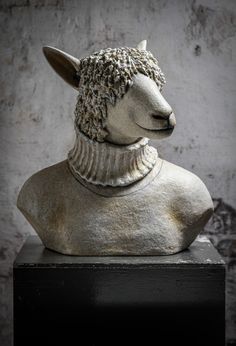 a statue of a sheep wearing a sweater on top of a black block in front of a wall