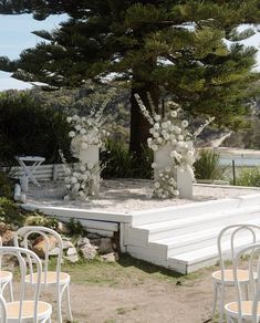 an outdoor ceremony setup with white chairs and flowers on the stage, surrounded by pine trees