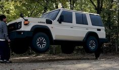 a man standing next to a white suv in the middle of a forest with trees