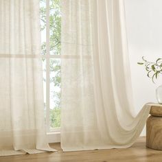 a white curtain with sheer drapes in front of a wooden table and vase filled with flowers