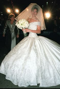 a woman in a white wedding dress holding a bouquet and walking down the street at night