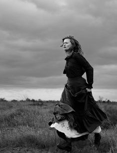 black and white photograph of woman in long dress standing on grass with cloudy sky behind her