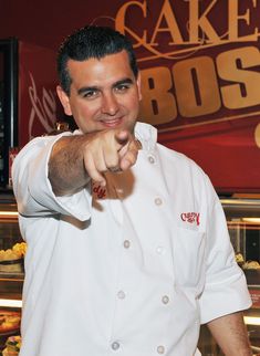 a man pointing at the camera while standing in front of a cake boss sign and display case