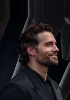 a man with long hair and beard wearing a black suit looks off to the side