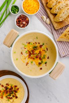 a bowl of potato soup on a table with bread, cheese and green onions next to it