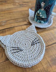 two crocheted cat coasters sitting on top of a wooden floor next to a coffee cup