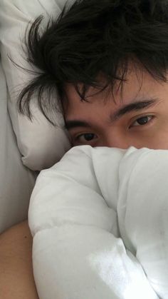 a young man laying in bed with white sheets and pillows on his head, looking at the camera