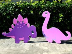 two pink and purple dinosaurs standing next to each other in front of some shrubbery