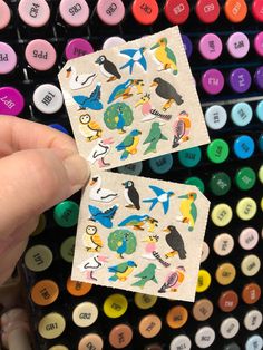 someone is holding up two small stickers with different colored birds on them next to some crayons