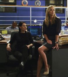 two women sitting at a desk in an office talking to each other and one man standing next to her