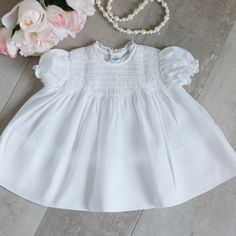 This Gorgeous White Smocked Baby Dress Is Heirloom Quality By Feltman Brothers. The Dress Is An All White Base. The Bodice Has Hand-Embroidered Pink And Green Rosebuds Atop The Smocking. Short Puff Sleeves Gather With Elastic And Have A Ruffled Hem. The Back Has A Yoke With A 3 Button Closure With Petite Pearlized White Buttons. The Neckline Is A Simple Rounded Shape With White Piping Trim. The Front Center Of The Bodice Has 3 Decorative Petite Pearlized Buttons Accented With Pink Embroidery Thr Feltman Brothers, Newborn Dress, Pink Embroidery, Newborn Dresses, Smock Dress, All White, Rose Buds, Kids' Dresses, Puff Sleeves