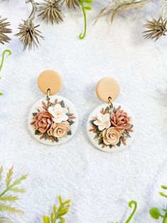 two pairs of earrings with flowers on them sitting in front of some pine cones and plants