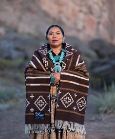 a native american woman wearing a brown blanket and turquoise necklace standing in front of a rock formation