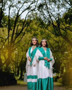 two women dressed in green and white standing next to each other on a path surrounded by trees