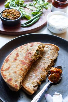 two flat breads on a black plate with spoon and sauce next to it, along with other food items