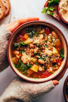 two hands holding a bowl of soup with beans and spinach on the table next to bread