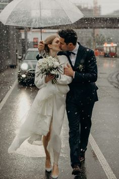 a bride and groom walking in the rain with an umbrella over their heads as they kiss