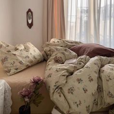 an unmade bed with flowers on it in front of a window and curtained windows