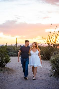 a man and woman walking down a dirt road at sunset with cactus in the background