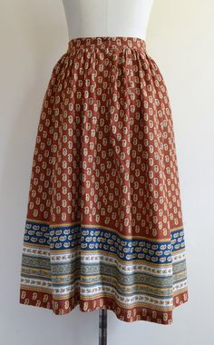 "This gorgeous skirt is gently gathered at the waist and comes in a handsome shade of rust--somewhere at the nexus of brown, red, and orange. The print features golden yellow paisley-like motifs as well as ornate bands of white and navy blue with pale green accents. Zippered back closure (nylon coil teeth) with an attractive plastic button. Good, wearable vintage condition. One tiny hole on the front left about the size of a pinhead (pictured), which appears to be a burn mark. We spotted two or Brown Vintage Tiered Skirt, Vintage Brown Tiered Skirt, Vintage Brown Flowy Skirt, Bohemian Brown Pleated Maxi Skirt, Traditional Long Brown Skirt, Vintage Long Brown Skirt, Vintage Long Skirt With Elastic Waistband, Spring Trench, Burn Mark