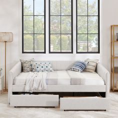 a white daybed sitting in front of two windows next to a book shelf with books on it