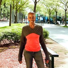 a woman standing next to a park bench in front of some trees and bushes, smiling at the camera