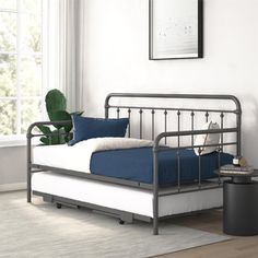 a metal day bed with blue sheets and pillows in a white room next to a potted plant