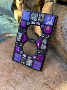 a purple and black mosaic tile light switch cover on a rock next to a body of water