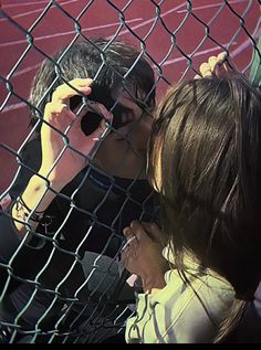 two people leaning against a fence talking on their cell phones while one person is holding a camera up to his ear