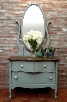 a blue dresser with flowers on top and a large mirror above the drawers, against a brick wall